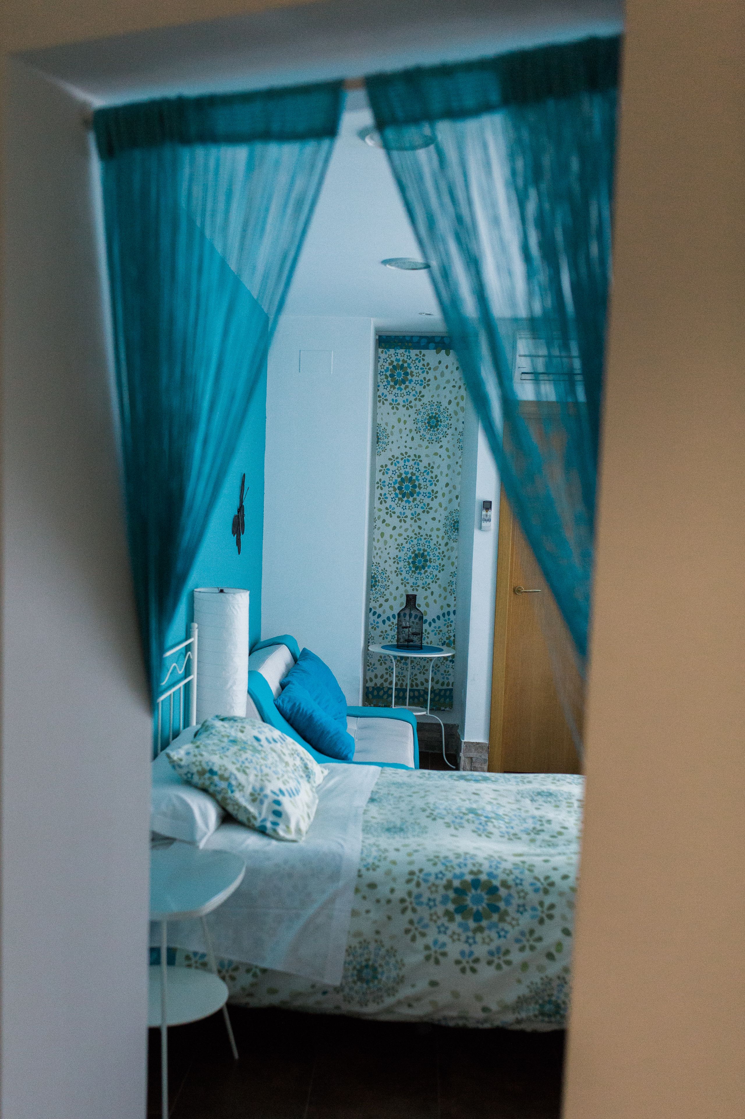 A bedroom with a bed, a bedside table, and a chair. There is a curtain partially drawn, revealing a bathroom with a toilet and a sink.