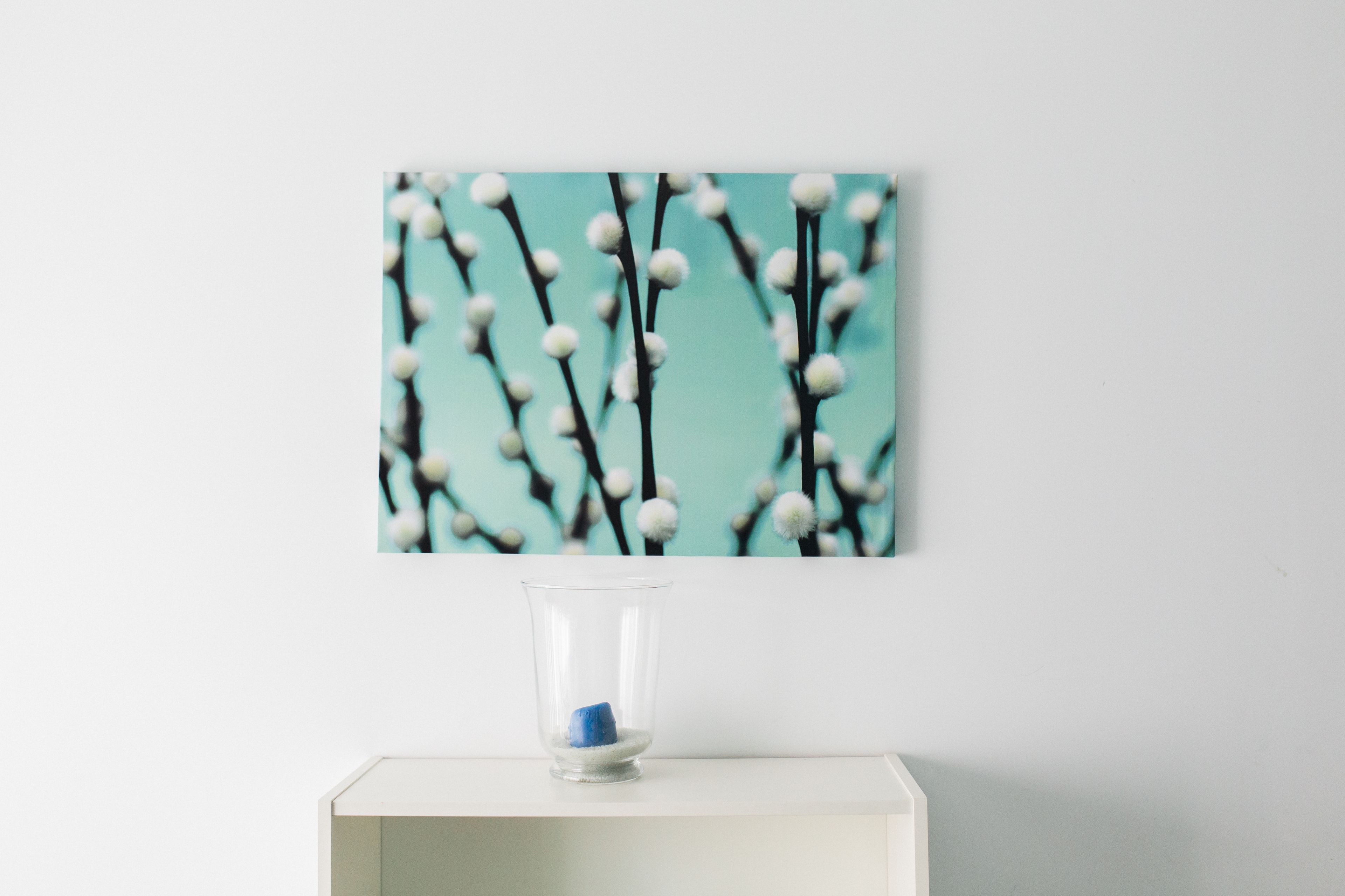 En genomskinlig glasvas med ett blått föremål i, placerad på en vit piedestal framför en vägg med ett botaniskt motiv.
