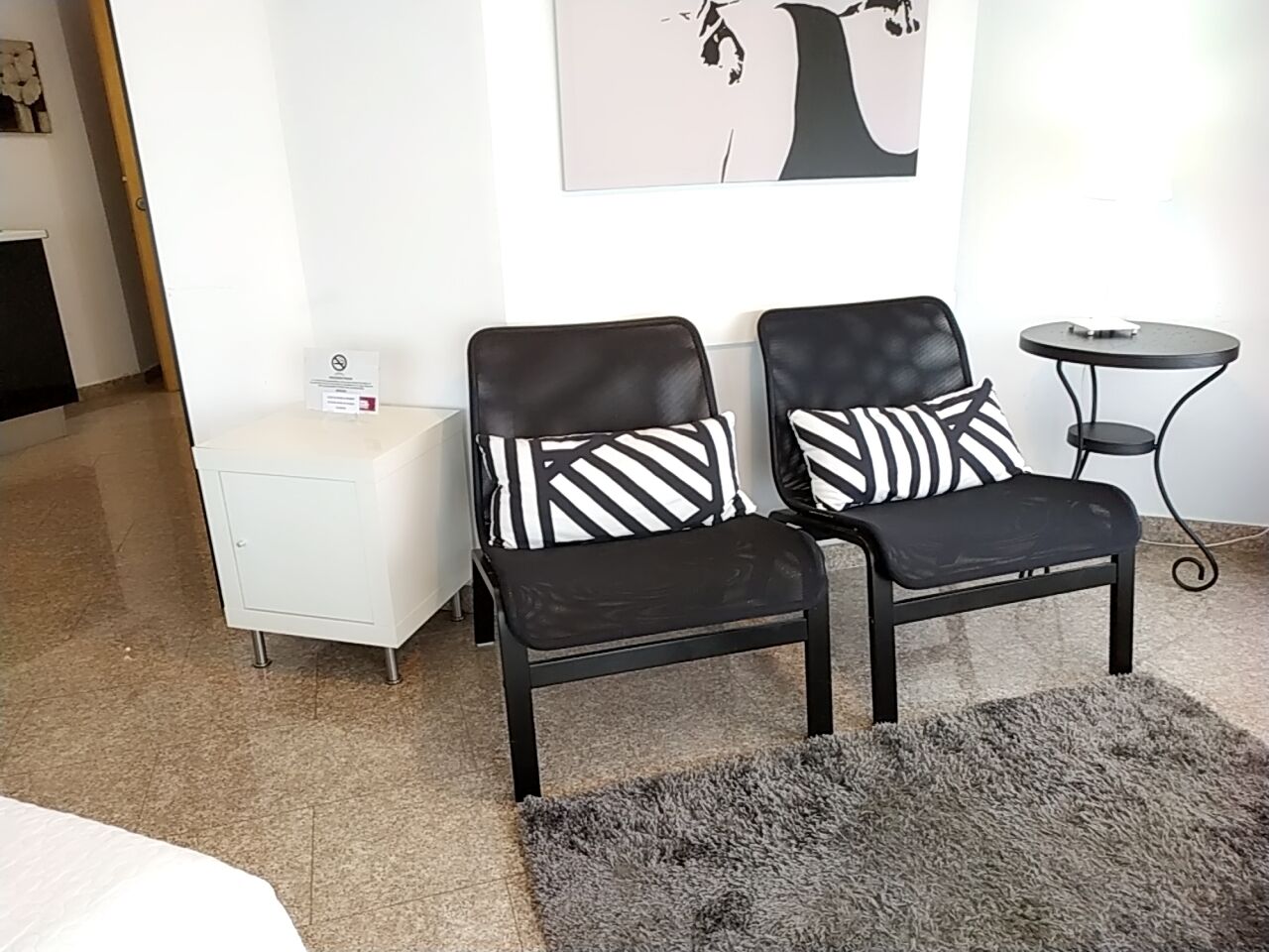 Et moderne hotelværelse med en seng, et hvidt natbord, to sorte stole med stribede puder, et lille rundt bord og en grå måtte.
