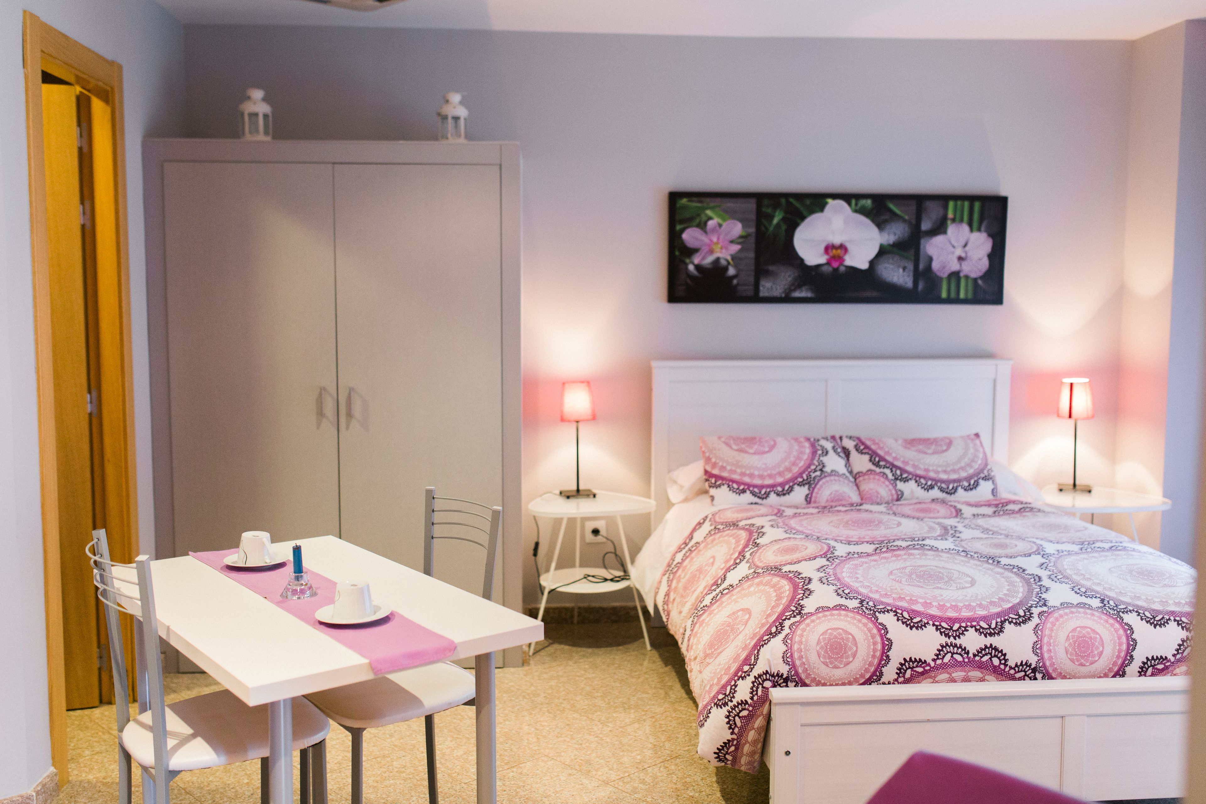 A bedroom with a bed, bedside tables, a desk, and chairs. There is a wardrobe and a painting on the wall.