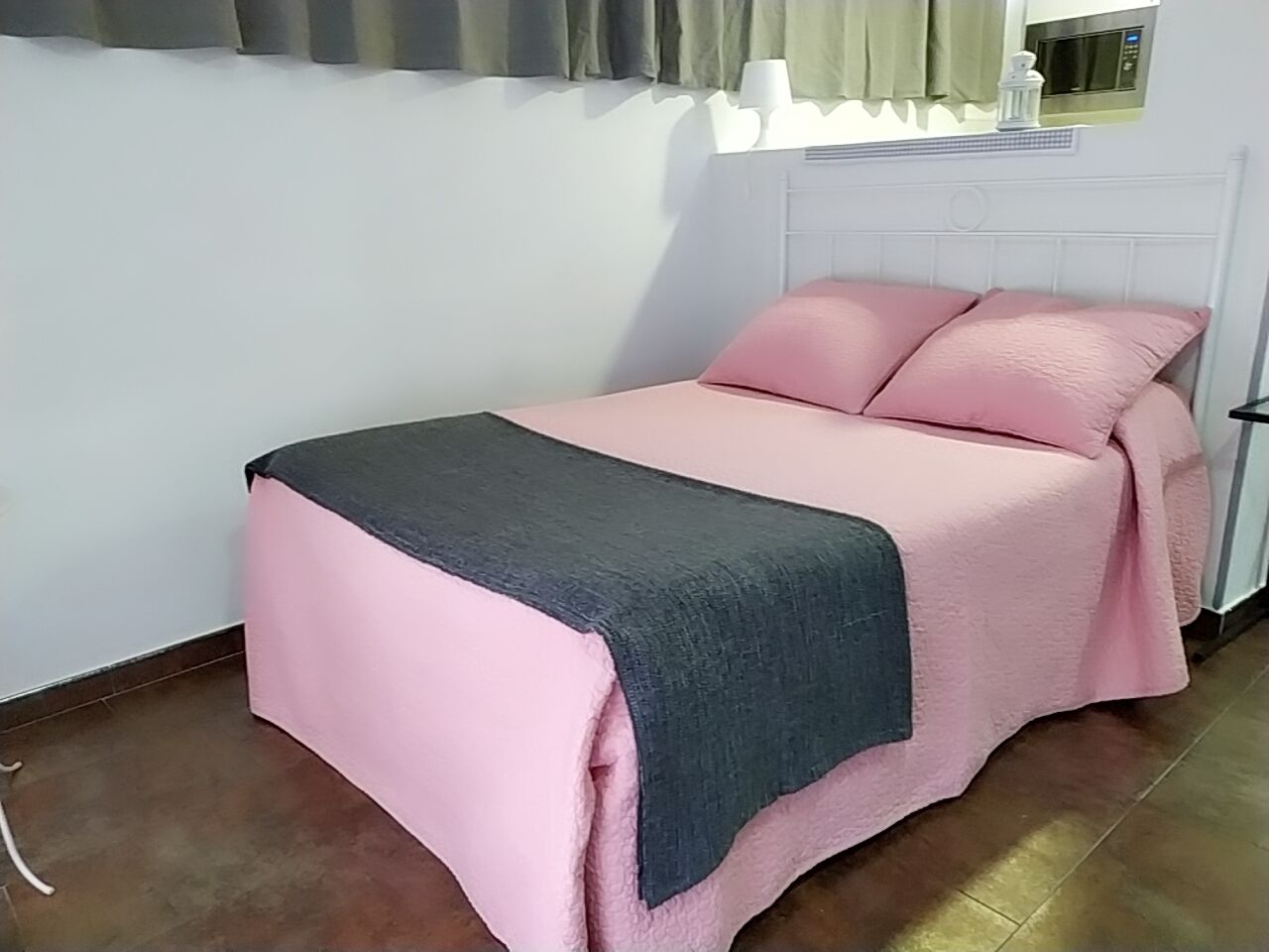 A single bed with a pink and grey bedspread, a white headboard, and a window with curtains.