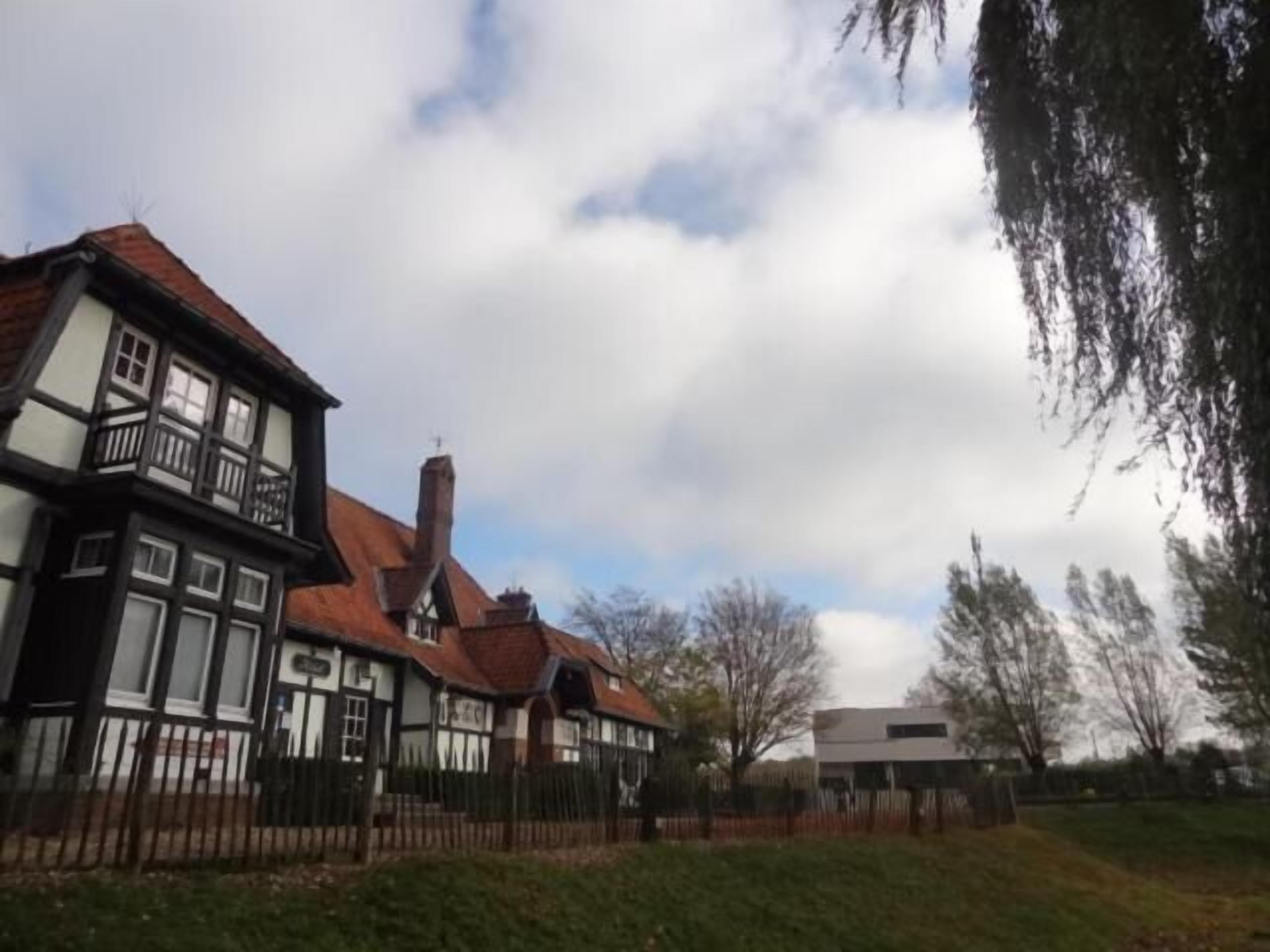 A traditional half-timbered house with a prominent chimney, surrounded by a fenced yard and a modern building in the background.