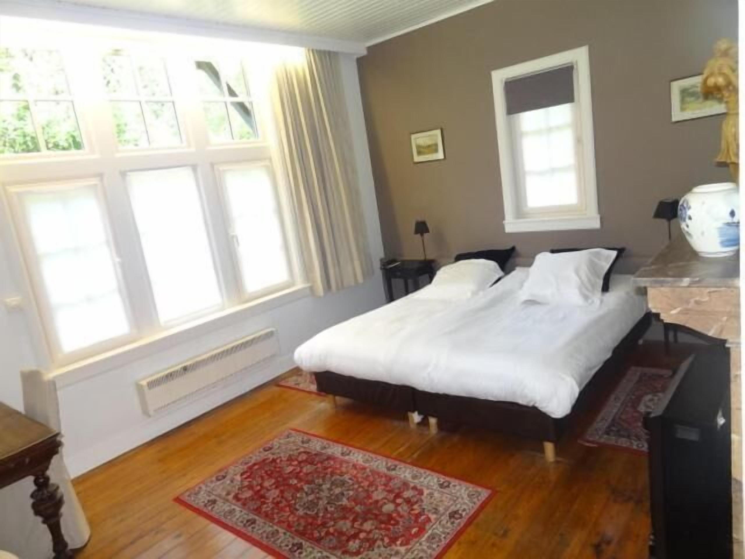 A bedroom with a bed, a window with curtains, a radiator, a nightstand, and a bedside table.