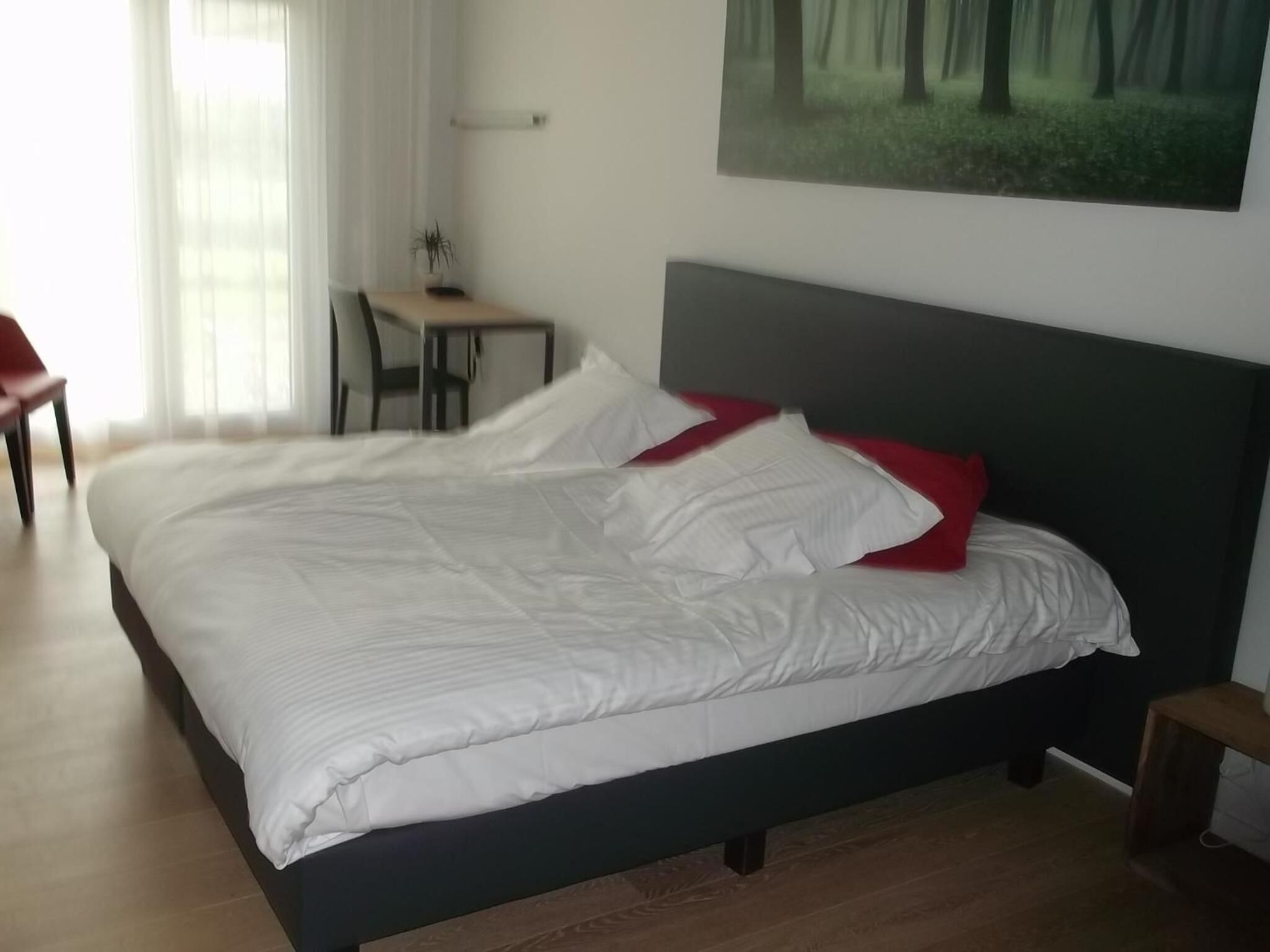 Un lit bien fait, avec une literie blanche et des coussins rouges, un petit bureau avec une chaise, et un tableau représentant une forêt accroché au mur.