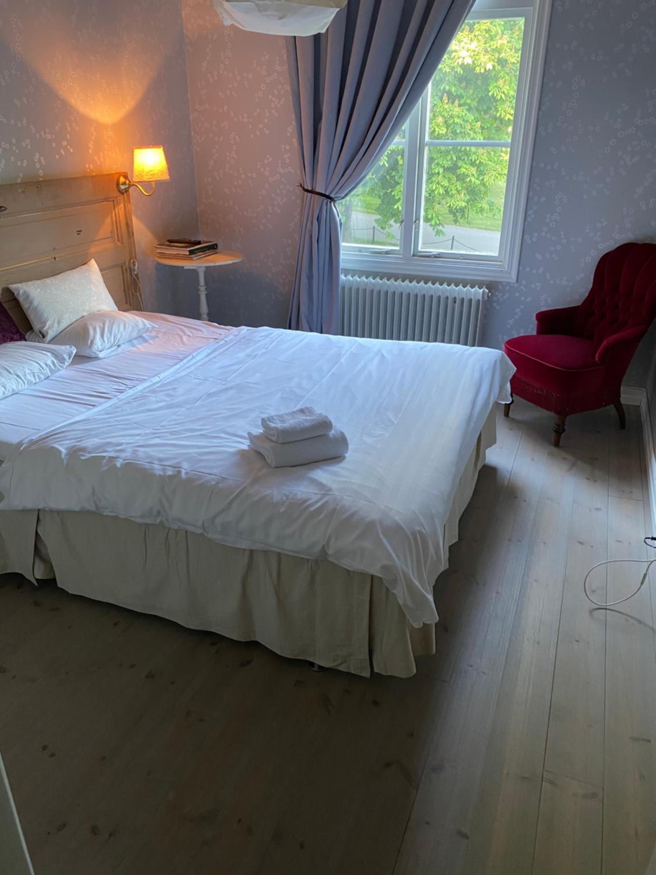 A neatly made bed with white linens, a red chair, a bedside lamp, and a window with curtains.