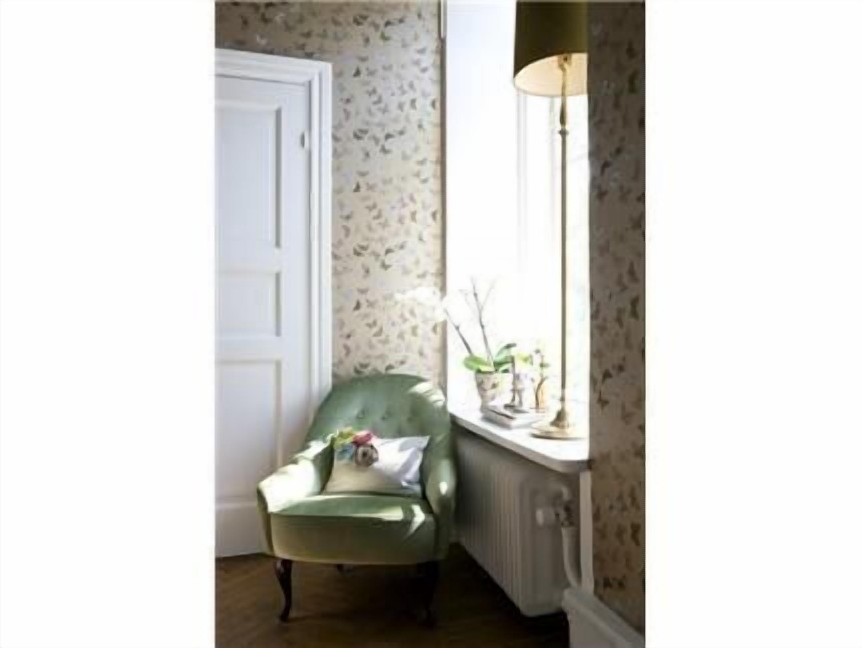 Una habitación de esquina con un sillón verde, una puerta blanca, un radiador y una ventana con una lámpara.