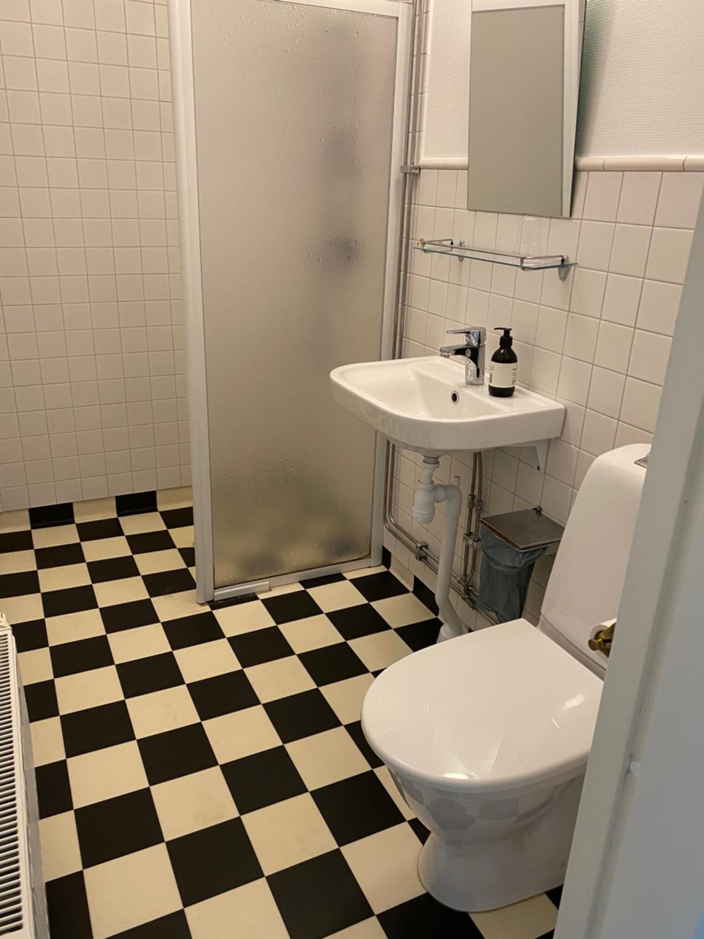 Un baño con piso a cuadros blanco y negro, un lavamanos blanco con un espejo arriba y un inodoro blanco.