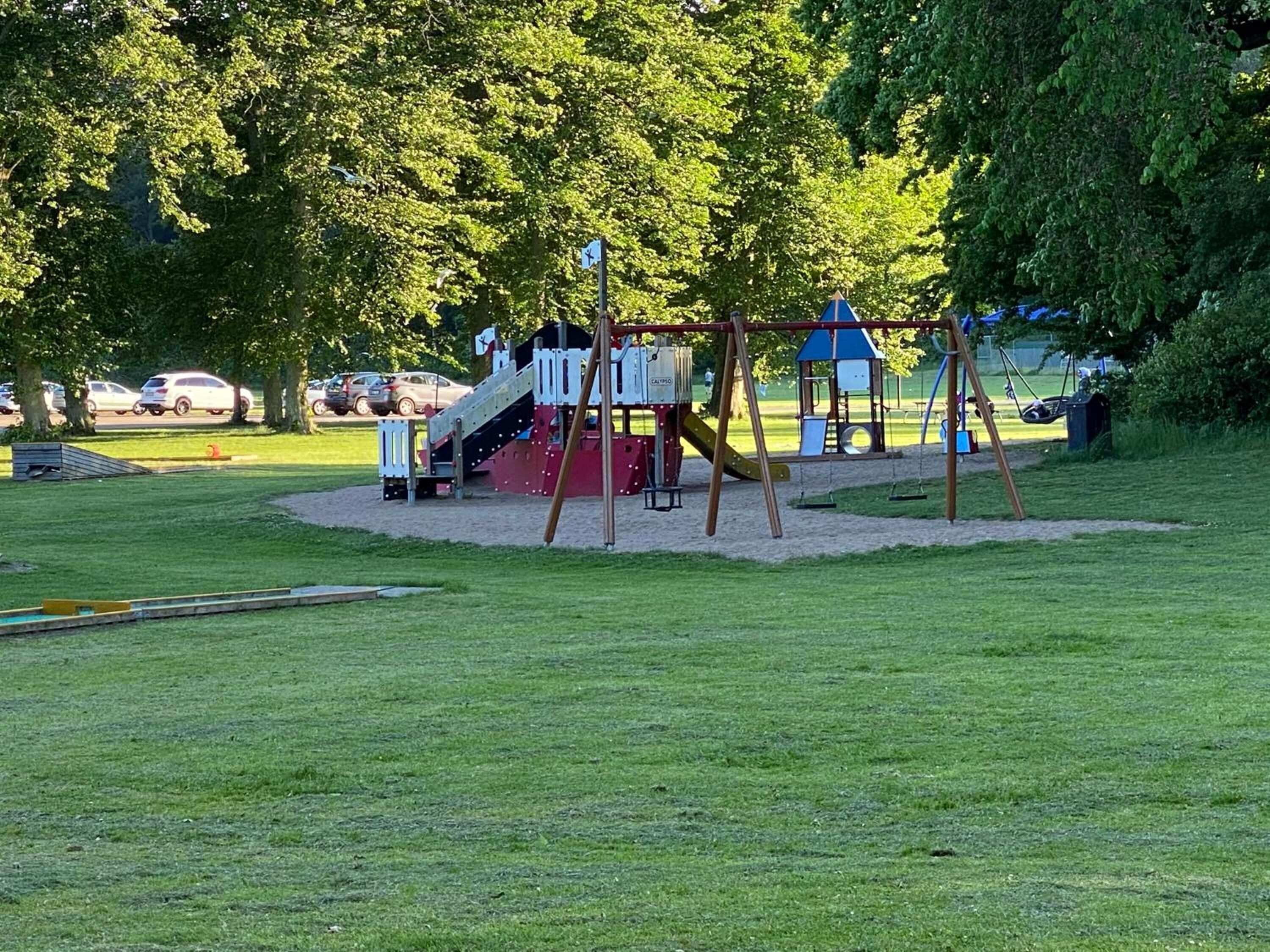Un parque infantil con tobogán y columpios, rodeado de árboles y un aparcamiento con coches.