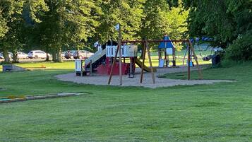 Un parque infantil con tobogán y columpios, rodeado de árboles y un aparcamiento con coches.