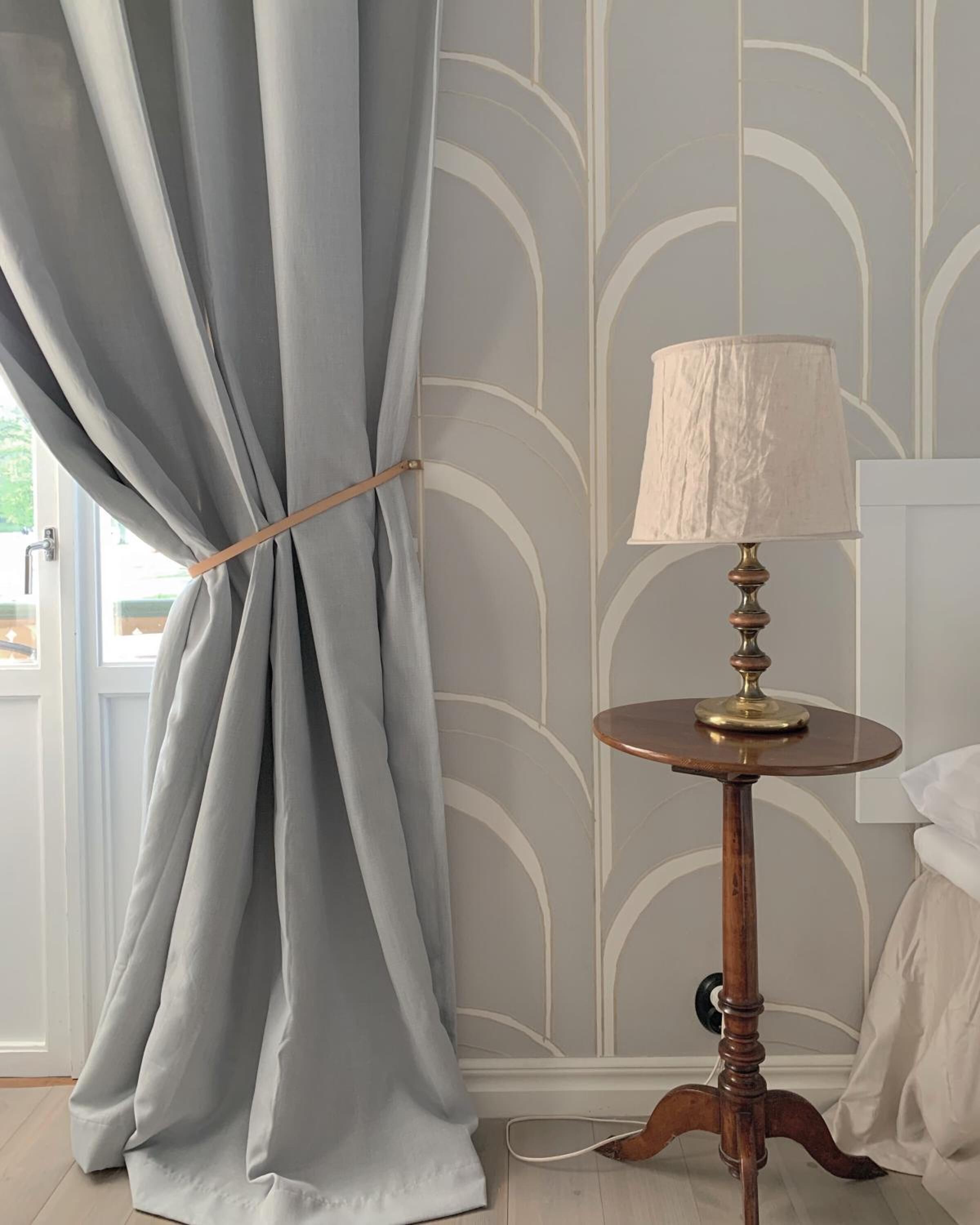 Una habitación con una ventana cubierta por cortinas azul claro sujetas con un lazo de madera, una mesa redonda de madera con base de latón y una pantalla blanca, además de una pared con un diseño geométrico.