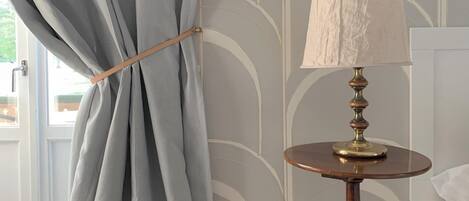 A room with a window covered by light blue curtains tied with a wooden tie, a round wooden table with a brass base and a white lampshade, and a wall with a geometric pattern.