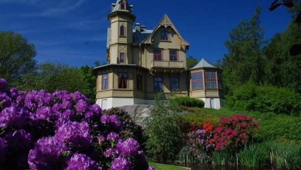 Una gran casa histórica con una torre destacada, rodeada de exuberantes jardines y un estanque.