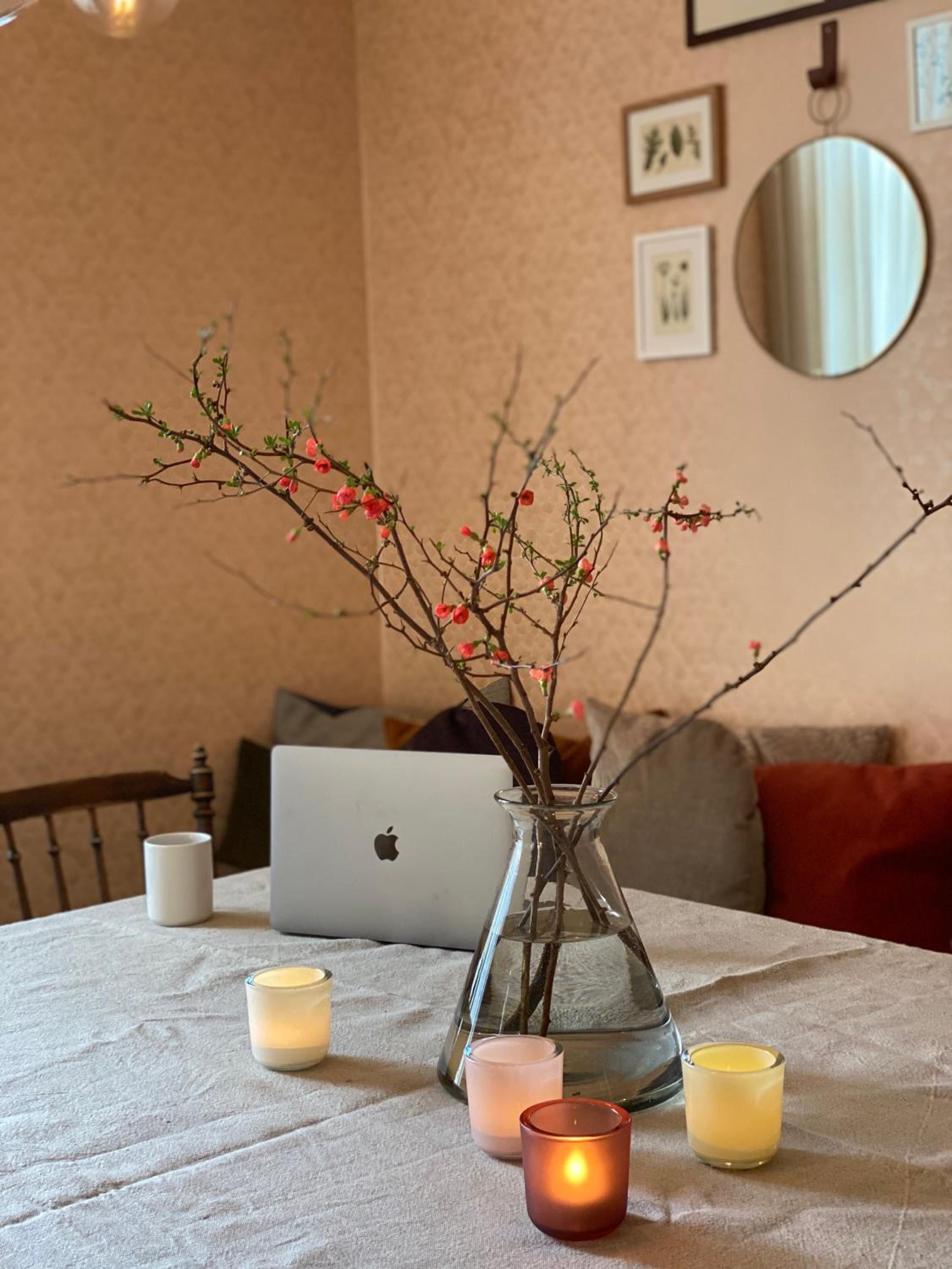 Una mesa con una computadora portátil, un florero con ramas y flores, y tres velas.