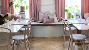 Un comedor con sillas y mesas de madera, un rincón con cojines junto a la ventana, y plantas en macetas.