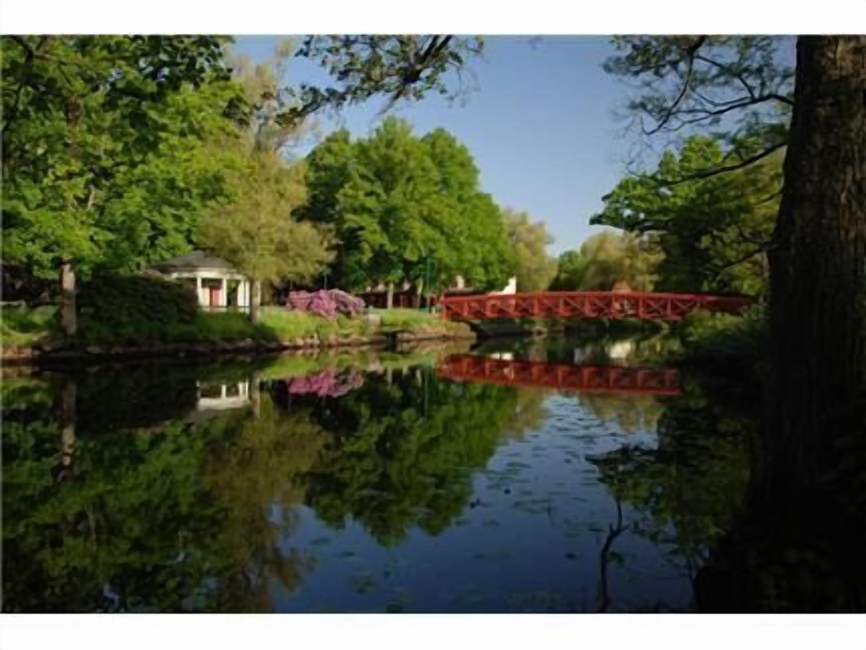 A serene riverside scene with a red bridge, lush greenery, and a small building with a red roof.