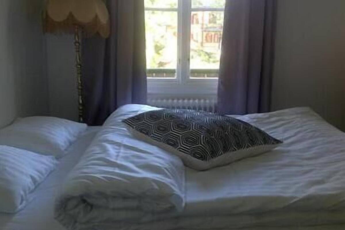 Ein ordentlich bezogenes Bett mit einem gemusterten Kopfkissen, ein Fenster mit Vorhängen und eine Stehlampe.