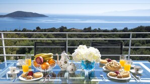 A table set with breakfast items and a vase of flowers, overlooking a scenic view of the sea and hills. - Villa Panorama (Agios Nikolaos)