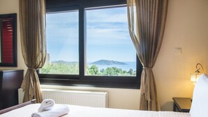 A hotel room with a bed, a window with curtains, a radiator, and a nightstand with a lamp. - Villa Panorama (Agios Nikolaos)