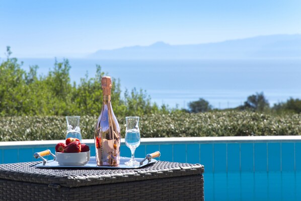 A table with a bottle, glasses, and a bowl of strawberries by a pool with a view of the sea and mountains.