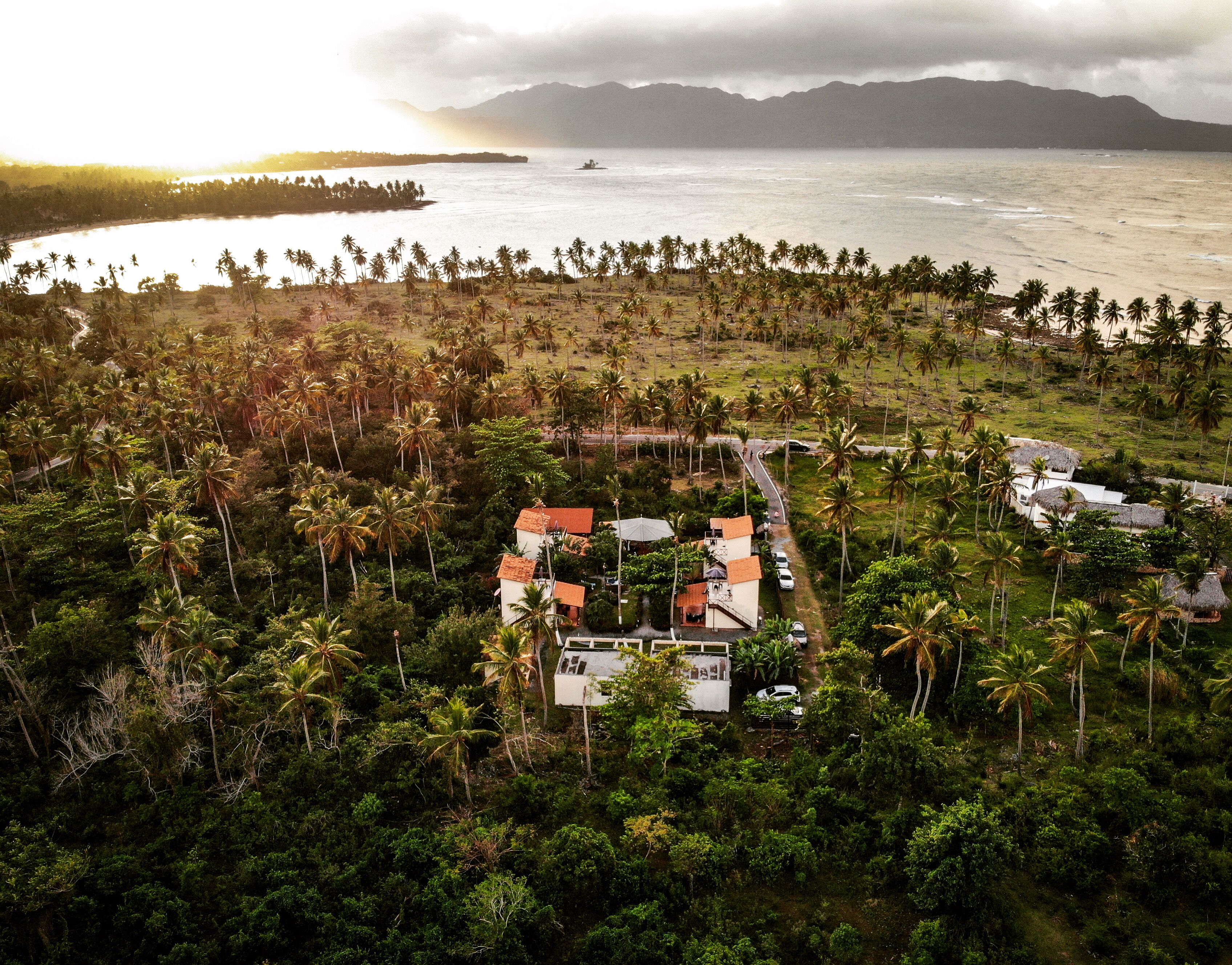 Vista aérea de una zona costera con un grupo de edificios rodeados de palmeras y una playa.