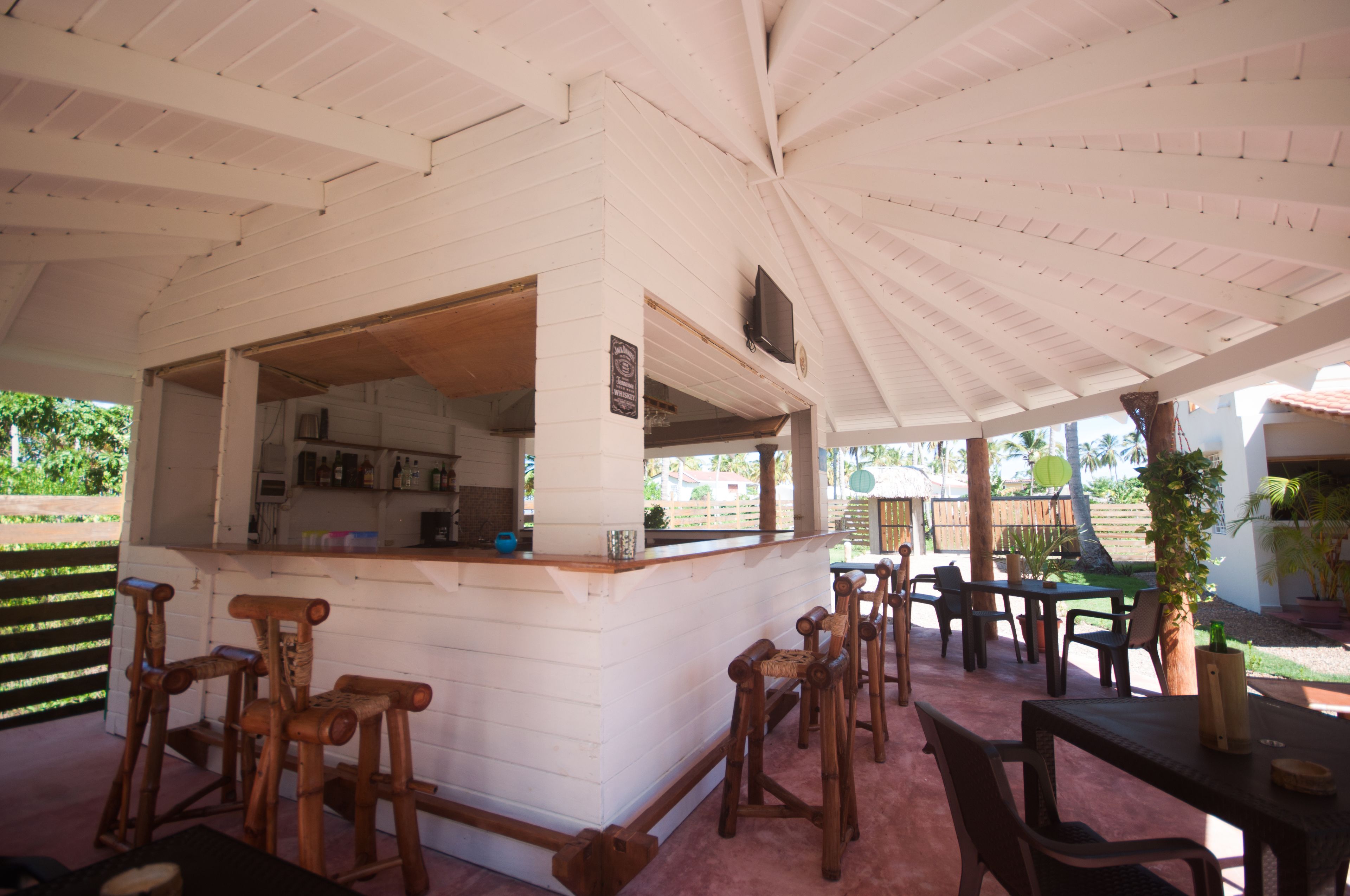 Un área de bar al aire libre, cubierta, con taburetes y mesas de madera, fachada blanca y vistas al jardín circundante.