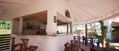 A covered outdoor bar area with wooden stools and tables, a white exterior, and a view of the surrounding garden.