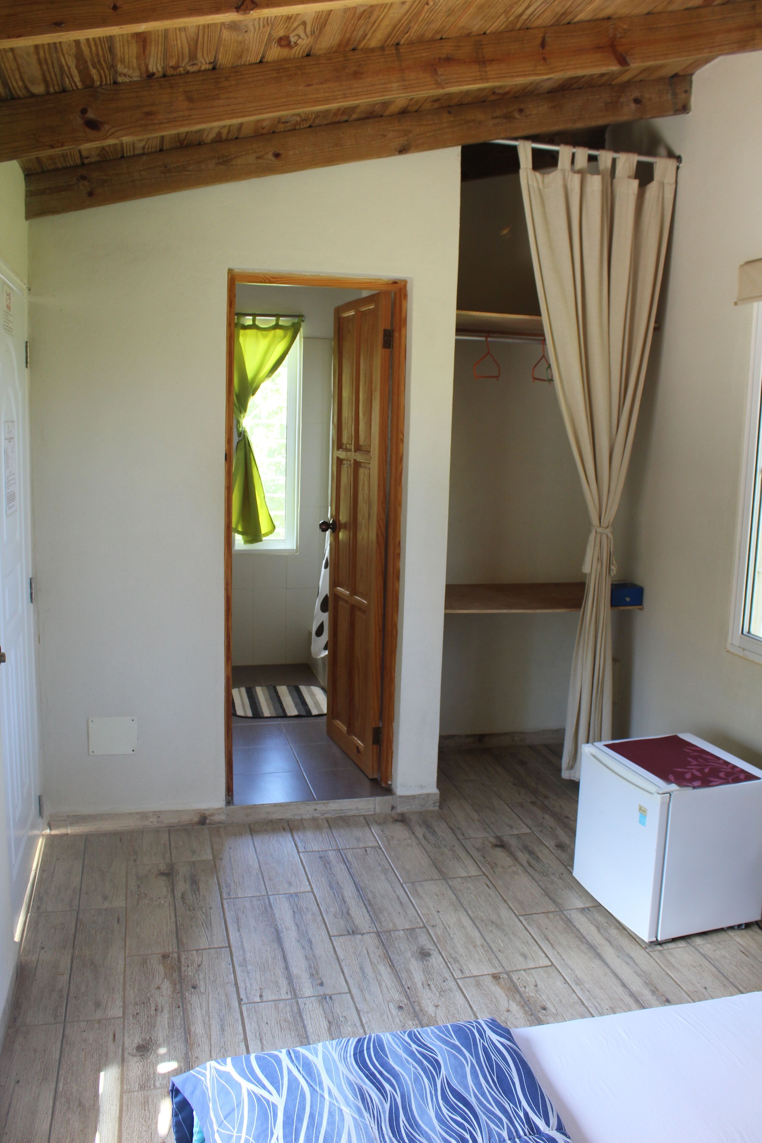 Une pièce avec un plancher en bois, un lit recouvert d’une courtepointe à motifs, un réfrigérateur blanc, une porte en bois et une fenêtre avec un rideau.