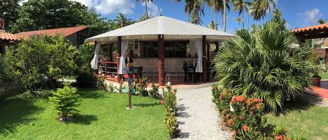 A covered outdoor seating area with tables and chairs, surrounded by lush greenery and palm trees.