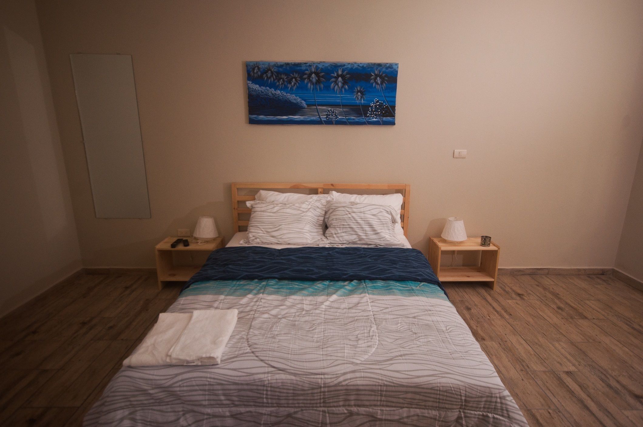 A neatly made bed with a patterned bedspread, two bedside tables with lamps, and a painting of palm trees on the wall.