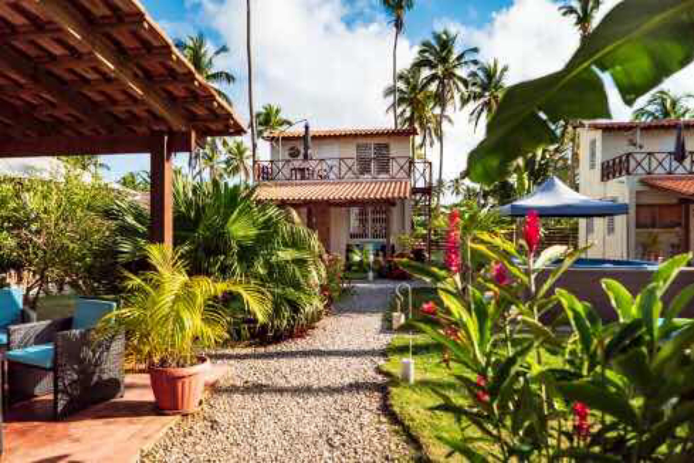 A hotel with a covered outdoor seating area, a pool, and lush tropical vegetation.