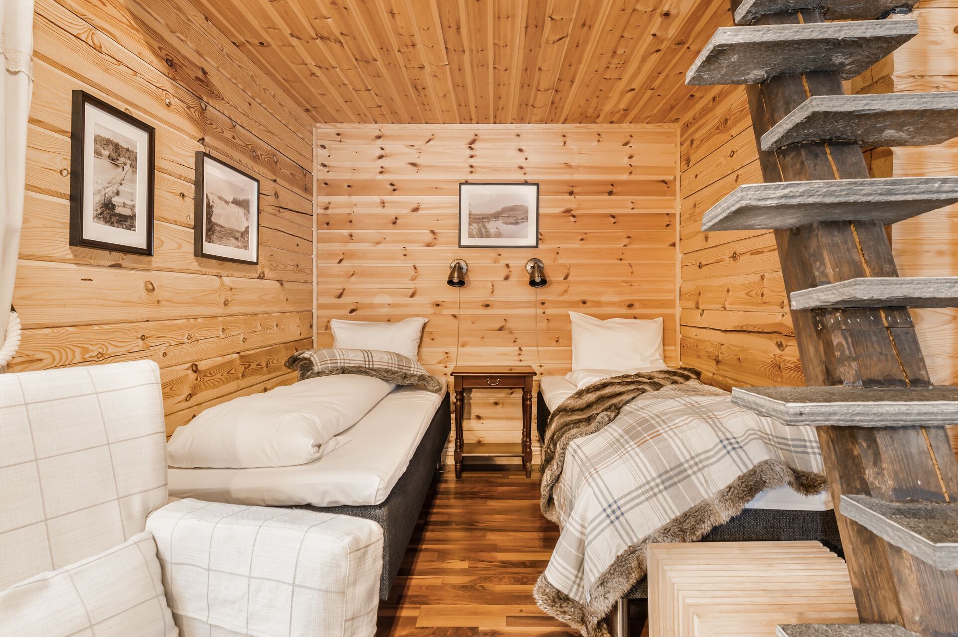 A cabin-style room with wooden walls and floors, two beds, a small table, and a wooden staircase.