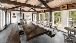 A spacious indoor area with a long wooden table, multiple chairs, and large windows offering views of the outdoors.
