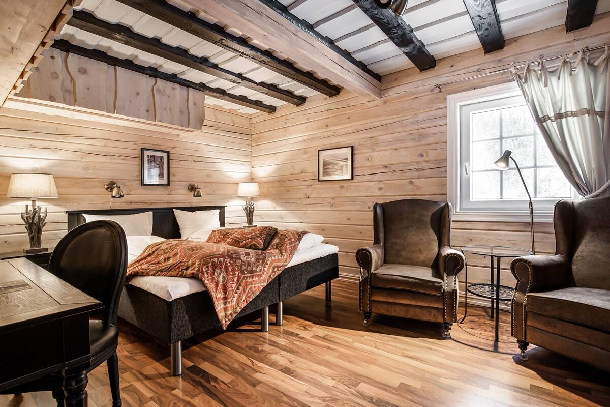 A log cabin-style room with a bed, two armchairs, a small table, and a window with curtains.