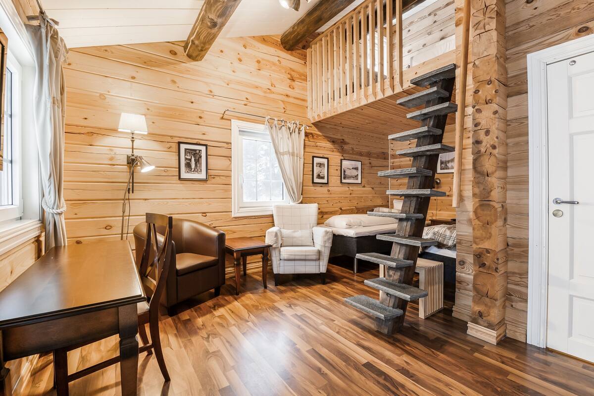 A wooden interior with a dining table, two chairs, a staircase, and a bed.