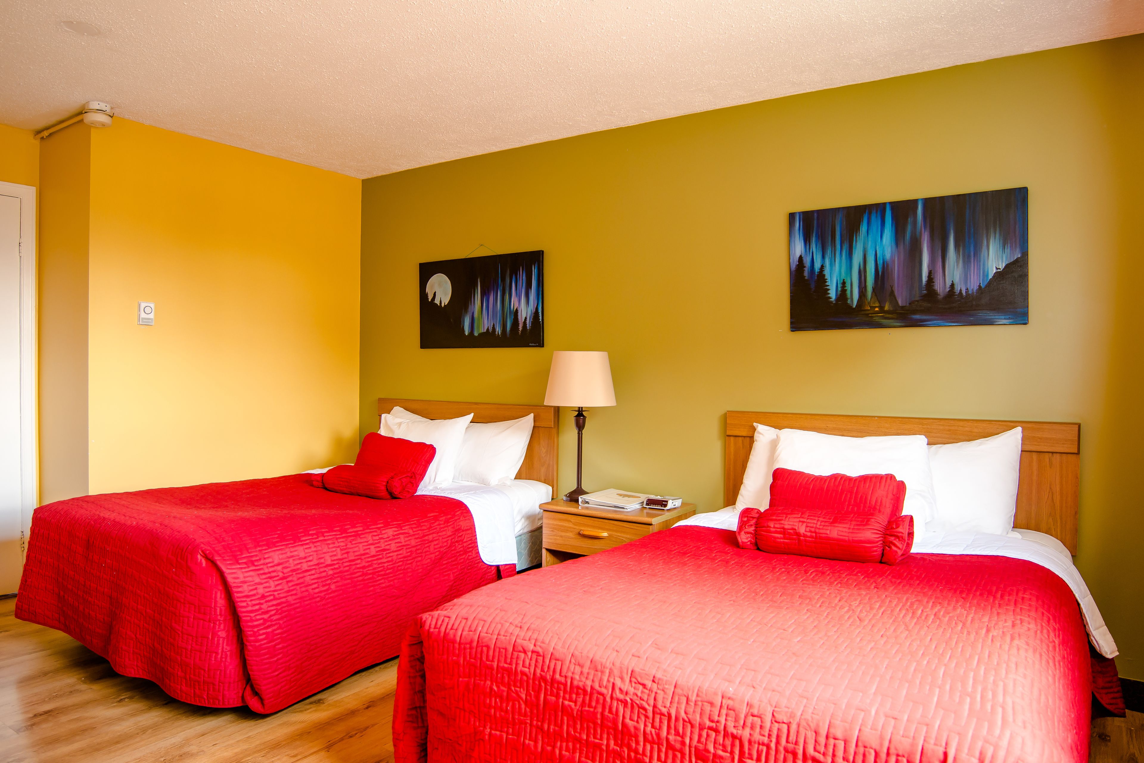 A hotel room with two beds, each with red bedding, a nightstand with a lamp, and two paintings on the walls.