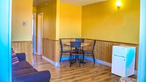Una habitación con piso de madera, una mesa de comedor con sillas, un refrigerador y un sofá con un cojín colorido.