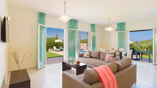 A spacious living area with a sofa, coffee table, and dining table. Large glass doors lead to a patio with outdoor seating and a pool. - Seven Springs Villas (Rhodes)