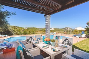 Un área de comedor al aire libre con vista a la piscina, muebles blancos y cielo despejado.