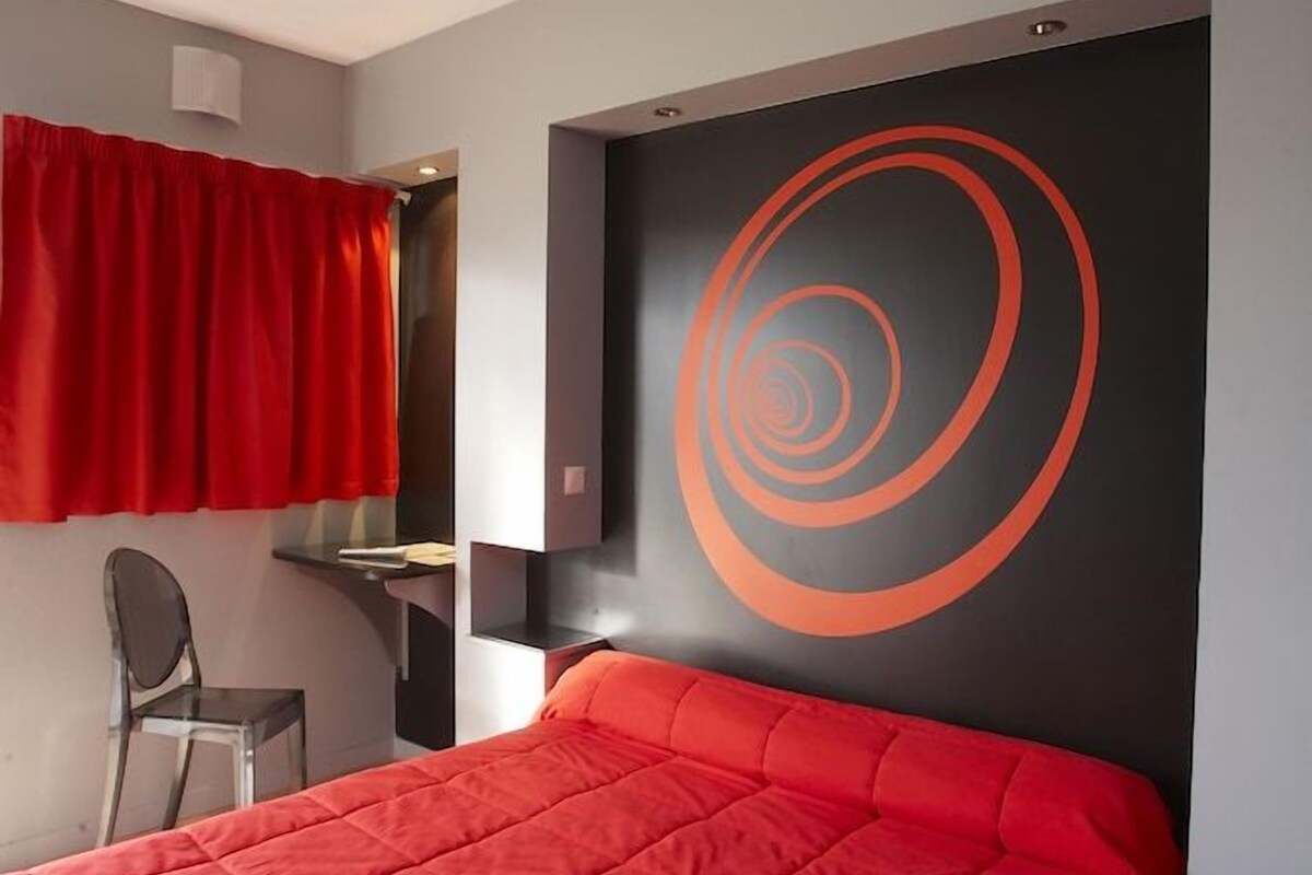 A bedroom with a bed, a chair, a desk, and a wall with a circular design.