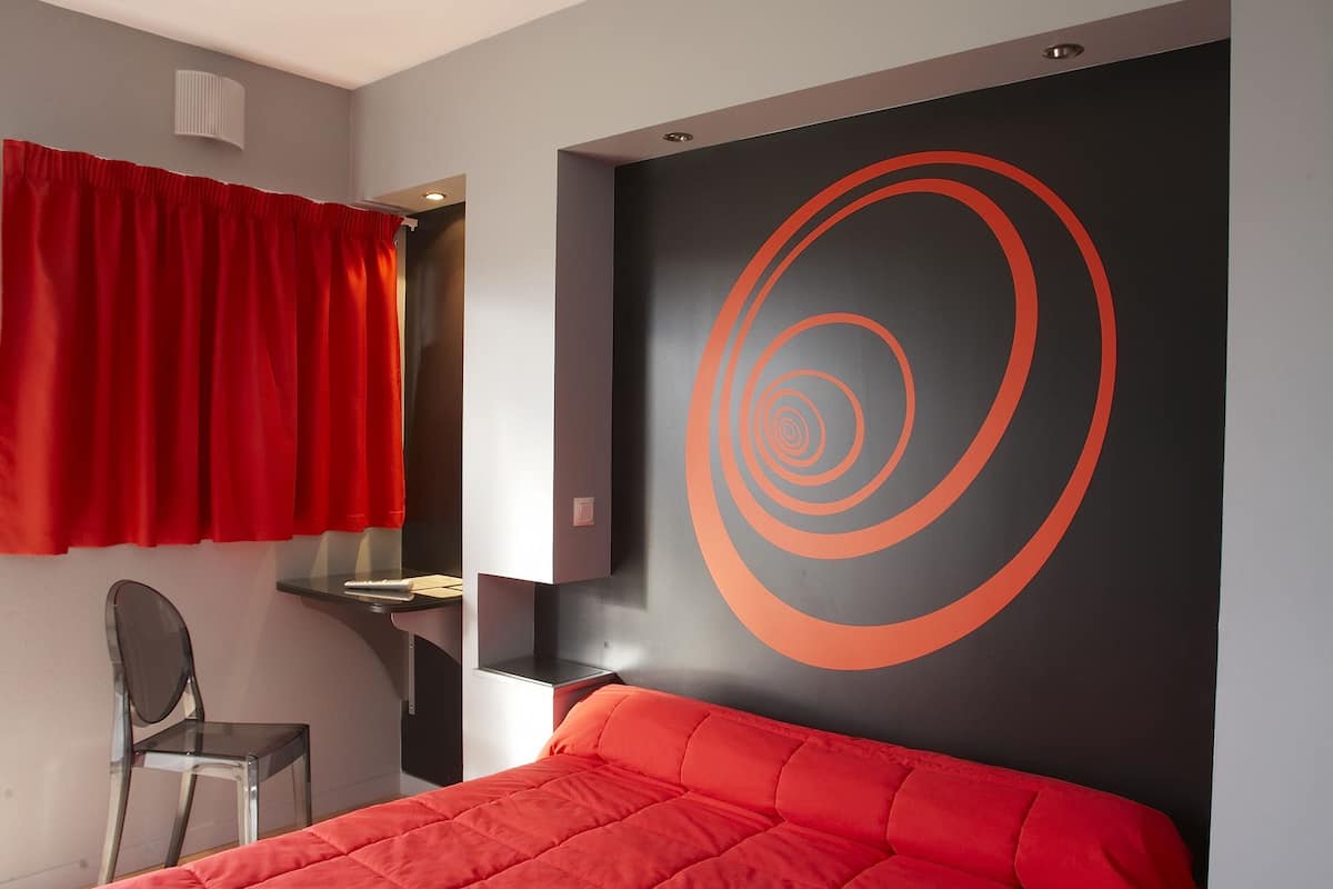 A room with a bed, a chair, a red curtain, and a wall with a circular design.