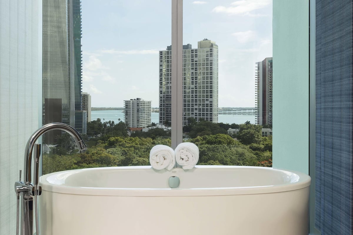Un baño con bañera, dos toallas y vistas a edificios altos y agua.