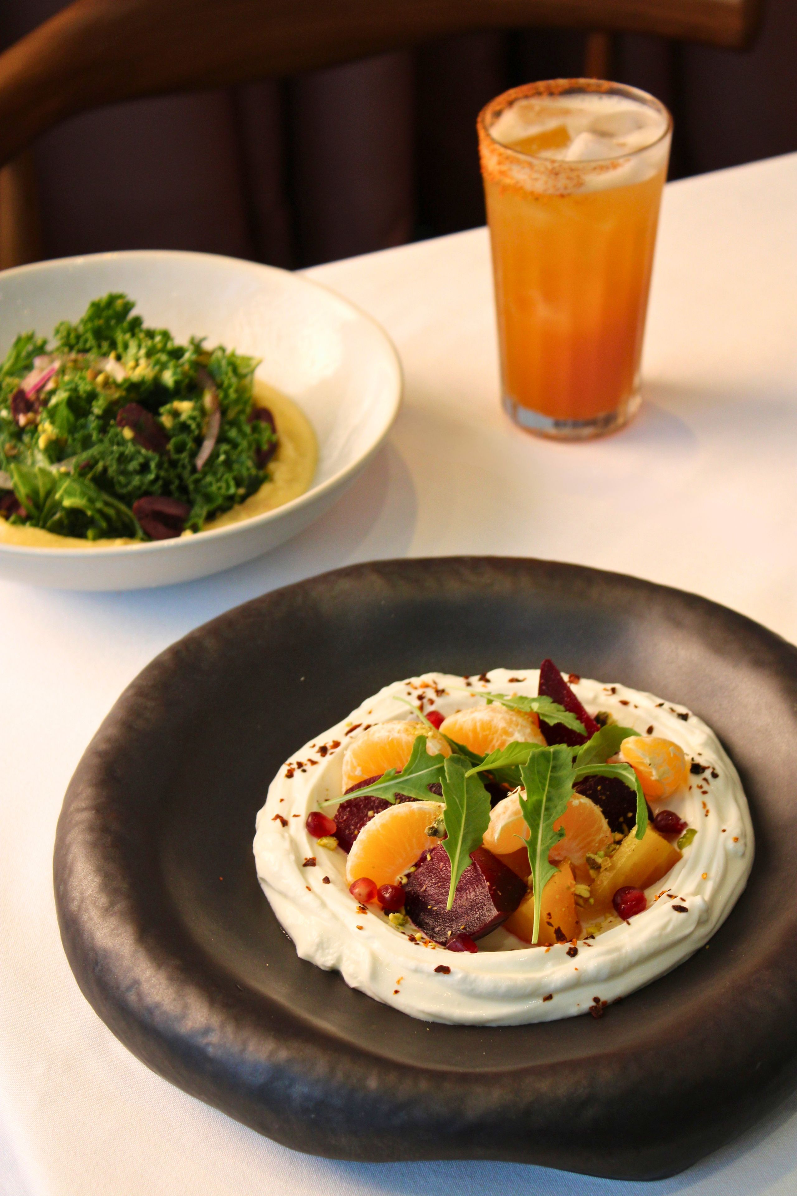 Une assiette de nourriture recouverte d’une sauce crémeuse et agrémentée de fruits et de légumes verts, un verre d’une boisson surmontée d’une mousse, et un saladier rempli de salade, de légumes verts et de noix.