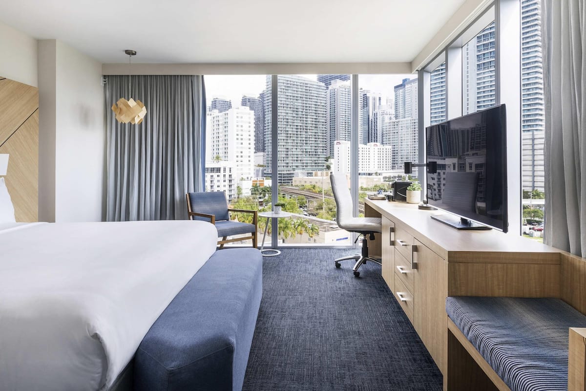 Habitación de hotel con cama, escritorio con televisor, una silla y vistas a edificios altos.
