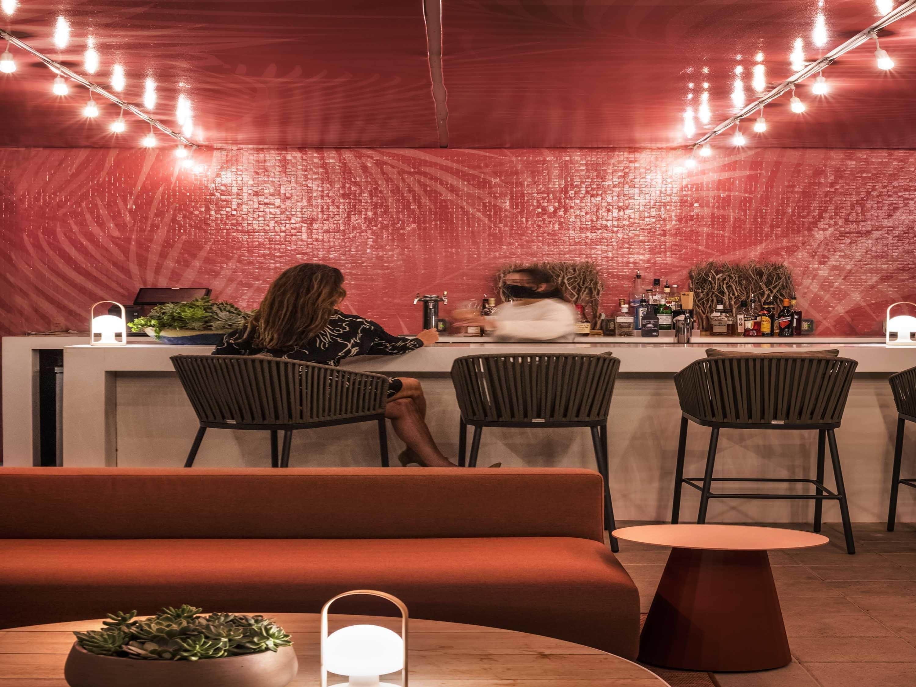 A bar area with a red textured wall, bar stools, a counter with a person behind it, and string lights.