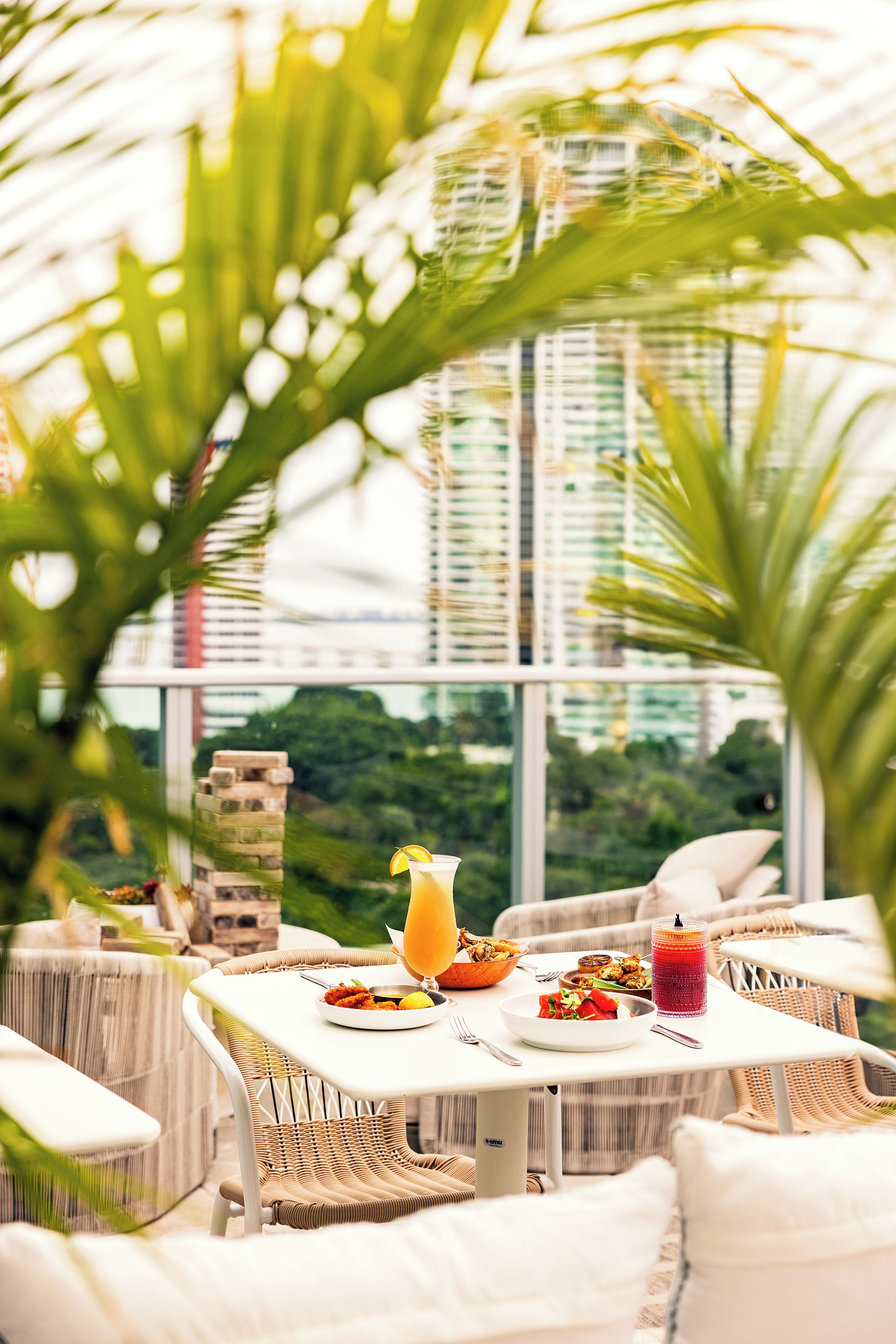 A dining area with outdoor seating, a table with food and drinks, and a view of greenery and buildings.