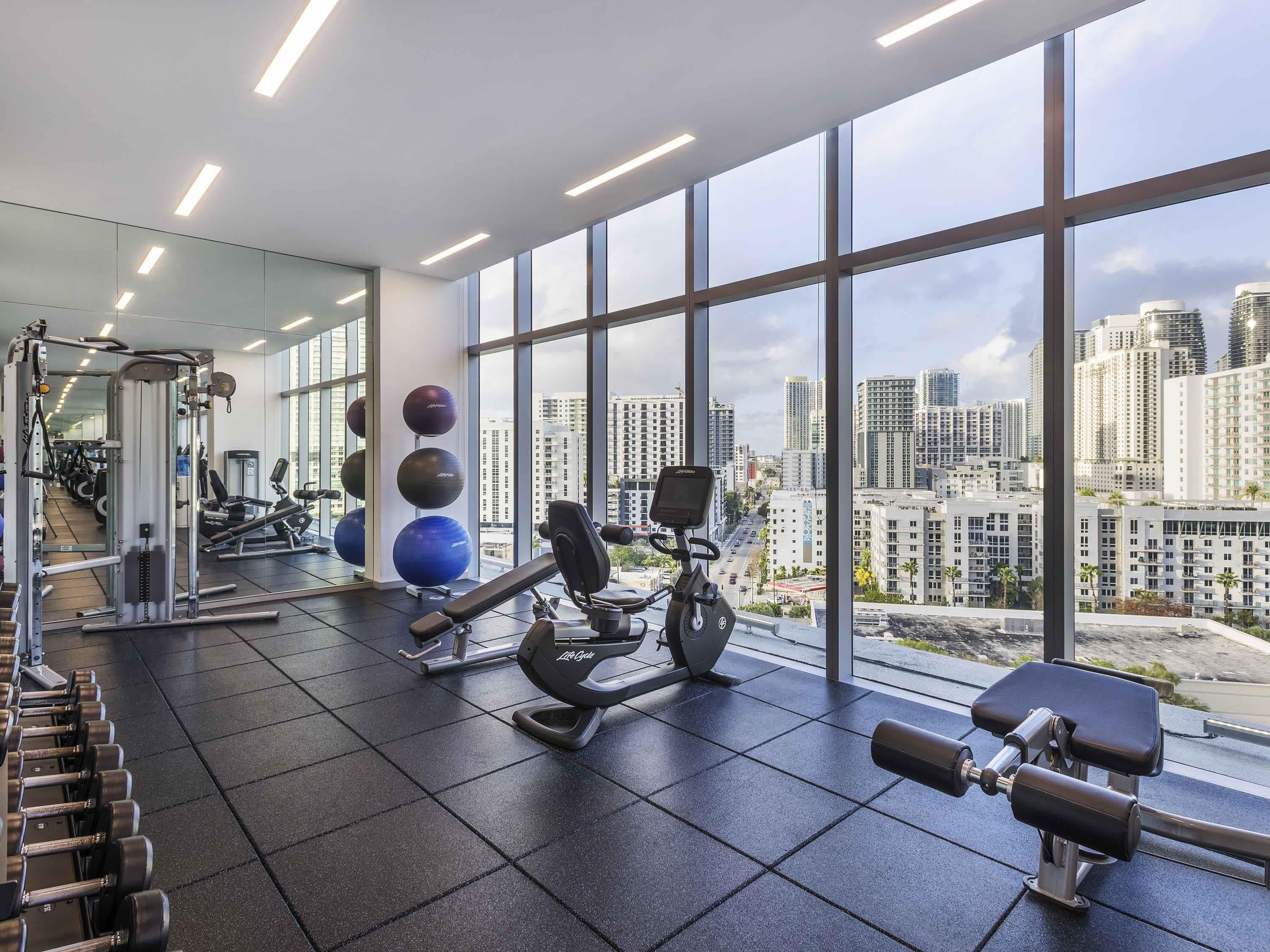 Un gimnasio con equipo de ejercicio, ventanas amplias y vista a la ciudad.
