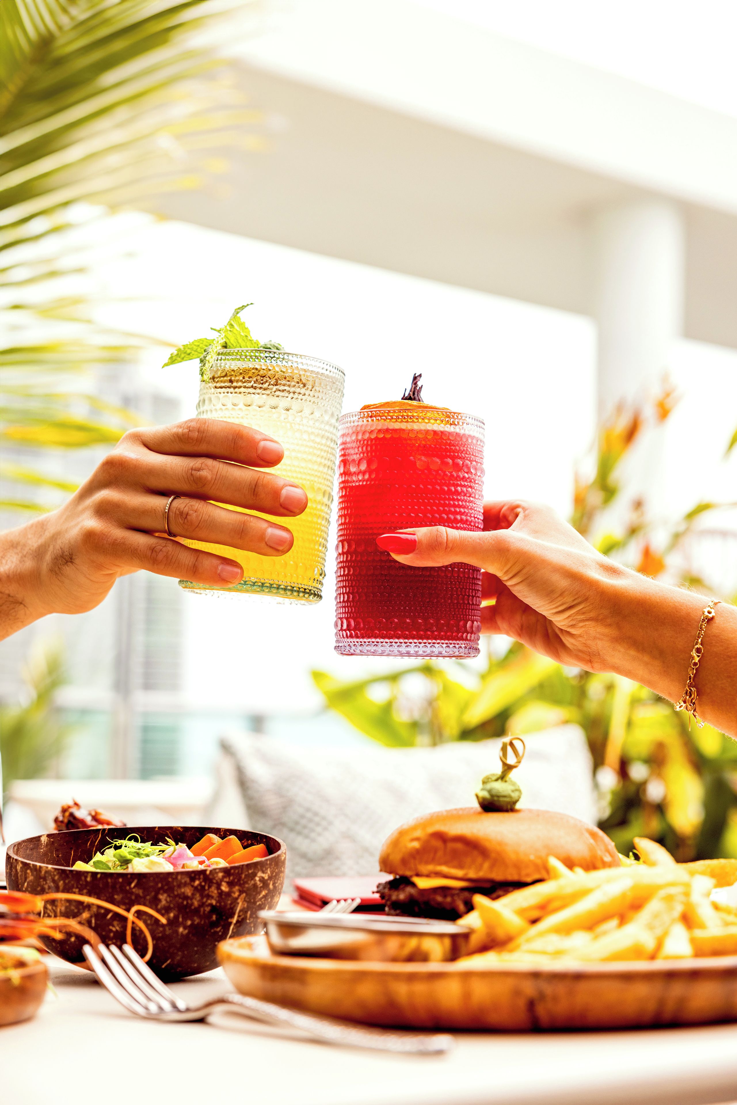 Dos manos sosteniendo vasos con bebidas, una hamburguesa, papas fritas y un bol de ensalada sobre una mesa.