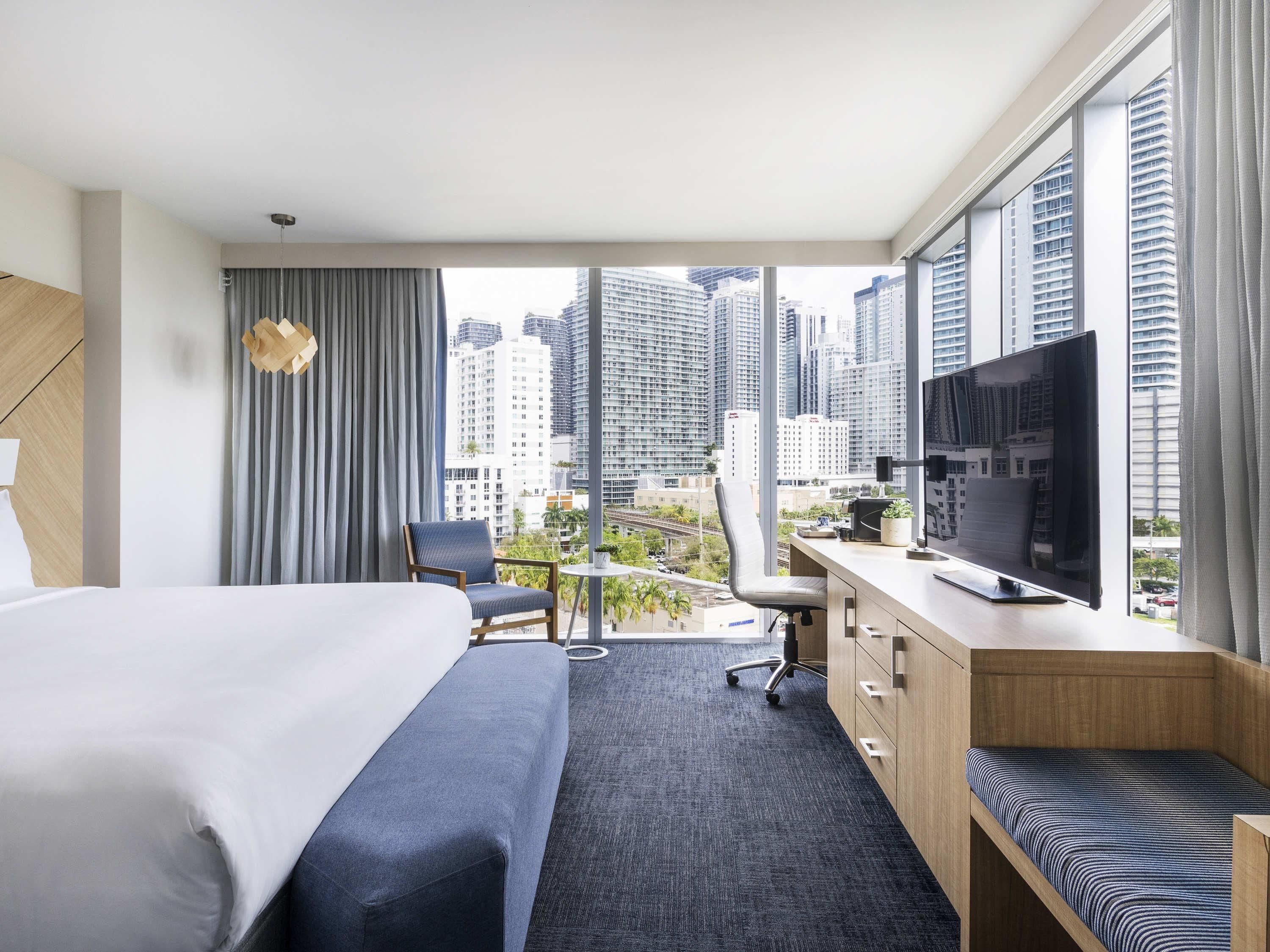 Habitación de hotel con cama, escritorio con televisor, una silla y vistas a edificios altos.
