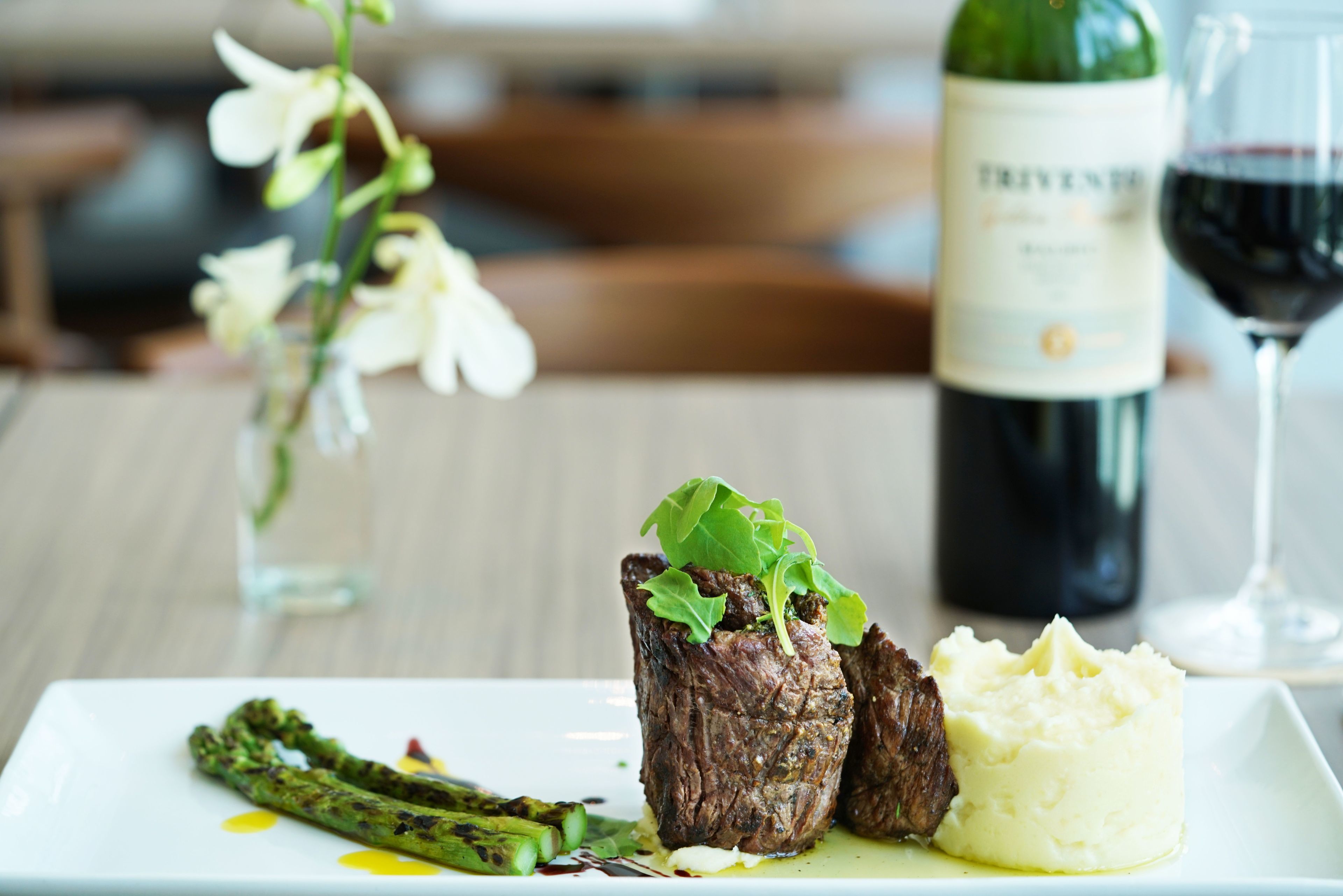 A plate with steak, asparagus, and mashed potatoes, a bottle of wine, and a glass of red wine on a table.