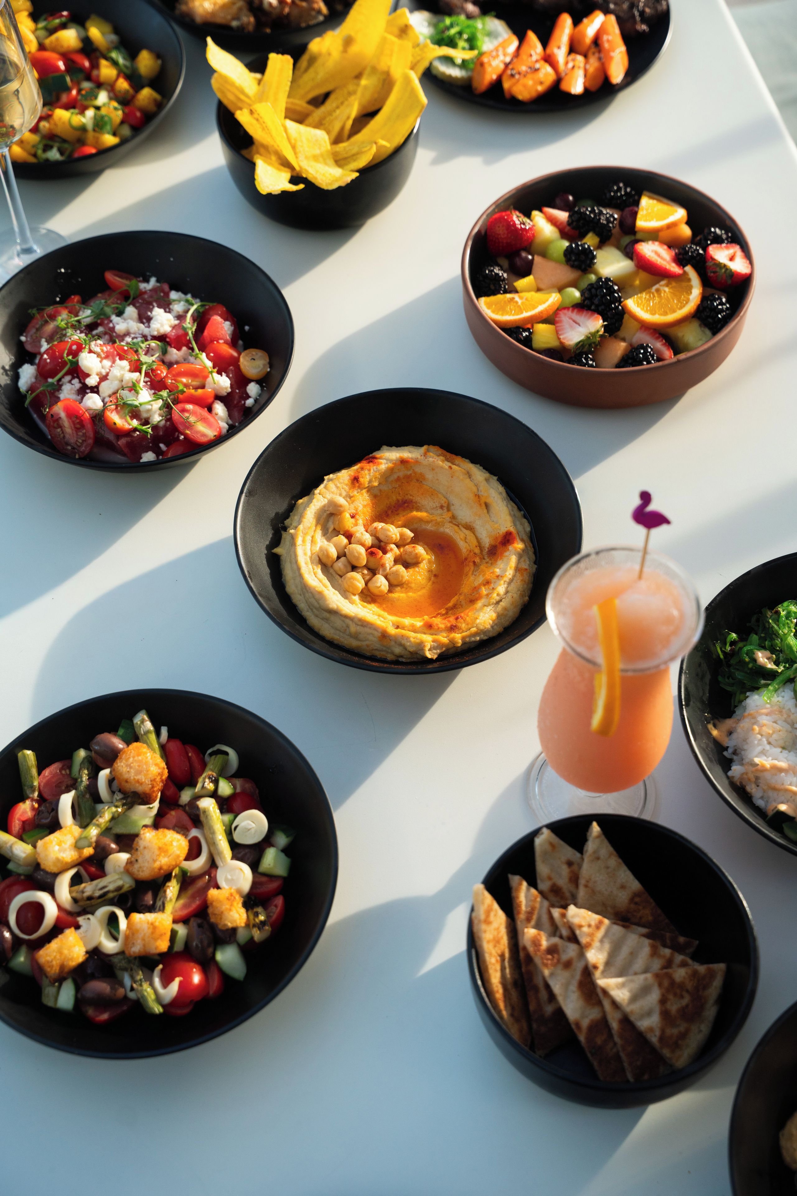 Ein Tisch mit verschiedenen Speisen, darunter Salat, Hummus, Obst und Brot.